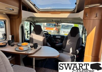 Interior of a camper van with a small dining table set with plates, croissants, and drinks; driver and passenger seats visible, skylight above; SWART CAMPERS logo bottom right.