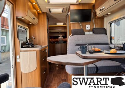 Interior of a camper van with wood cabinets, a kitchenette, and a dinette set with coffee, croissant, and glasses; bed area visible in back.