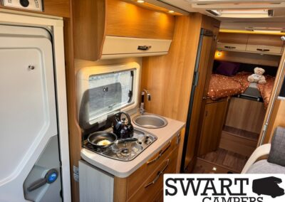 Campervan kitchen with a two-burner stove, kettle, and sink, wood cabinets, and a rear sleeping area with a teddy bear on the bed.
