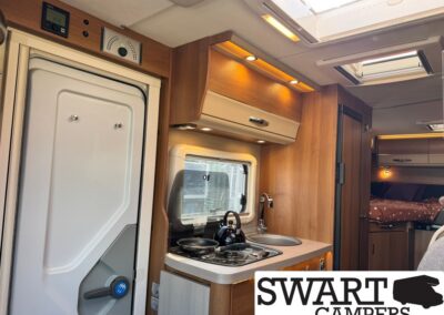 Interior of a camper van with a compact kitchenette: stove with kettle, sink, window, and wooden cabinets.