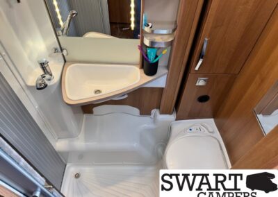 Compact camper bathroom with white sink, mirrored cabinet, toothbrush holder, and wooden storage cabinets; small shower base and a compact toilet visible; SWART CAMPERS logo in bottom right corner.