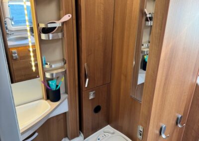 Small camper bathroom with wood cabinet, sink, mirror, and vertical storage shelves beside a compact toilet area. A Swart Campers logo is in the corner.