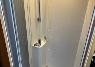 Compact white shower stall in a camper van with a chrome vertical shower rail and control knobs on the wall.