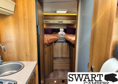 Cozy camper van interior with a kitchenette on the left and a bed area at the far end, a small teddy bear sits on the bed.
