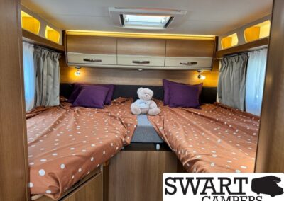 Twin camper bed setup with orange polka-dot sheets, purple pillows, and a teddy bear between the beds under overhead cabinets.