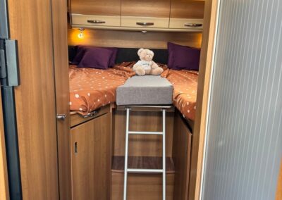 Camper bedroom with two single beds on either side, orange polka-dot bedding, purple pillows, a gray step stool with a teddy bear, overhead cabinets, and a skylight. SWART CAMPERS branding in corner.