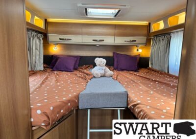 Cozy camper van bedroom with twin bunks, orange polka-dot sheets, purple pillows, a teddy bear, and overhead cabinets.