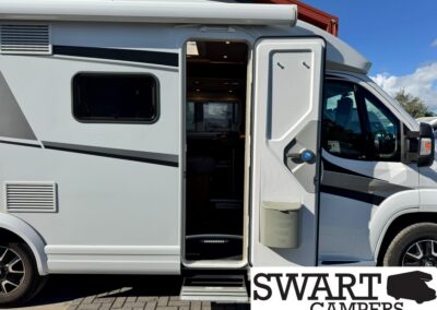 Side view of a white motorhome with gray and black graphics; the entry door is open, revealing the steps and interior.