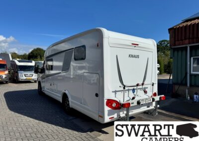 Rear view of a white KNAUS motorhome with a bike rack on the back, parked on a brick lot with other campers and a building nearby; SWART CAMPERS logo appears in the lower-right corner.