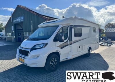 White motorhome parked on a paved lot beside a dark green service workshop bearing 'Garage Swant' (Blurry sign) in the background.