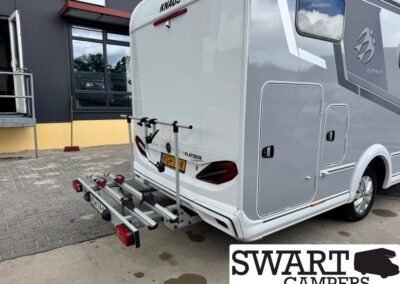 Rear view of a white KNAUS motorhome with a two-bike rack attached to the bumper, parked on a paved lot beside a dark building; SWART CAMPERS logo visible in the bottom-right.