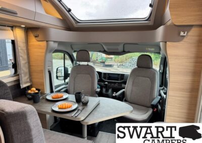 Camper van dining area with a table set for two croissants, cups, and cutlery, front seats in view and a large roof skylight above.