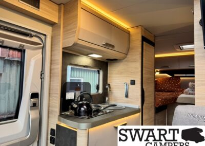 Interior of a compact camper van: small kitchen with kettle on stove, sink, and overhead cabinets; window, warm wood finishes, and a sleeping area with an orange bedspread and a teddy bear. Swart Campers logo visible in the corner.