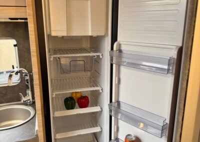 Open camper fridge with three peppers (green, yellow, red) on a shelf and a can of soda in the door pocket; watermark says SWART CAMPERS.