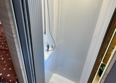 Compact white shower cubicle inside a camper, featuring a handheld shower on a vertical rail, chrome fixtures, built-in base, and two ceiling lights. A doorway leads into a sleeping area with polka-dotted bedding visible to the left.