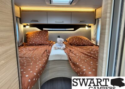 Compact camper bedroom with two single beds in rust-colored, white-polka-dot duvets, a teddy bear between them, and overhead cabinets with warm lighting.
