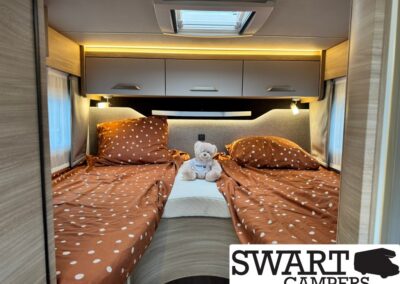 Two twin beds with rust-brown polka-dot bedding meet in a camper cabin, a teddy bear sits between them on a white mat.