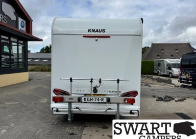 Rear view of a white Knaus motorhome with a bike rack, license plate KGH-74-R, parked near a camper dealership sign that says Swart Campers.