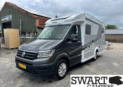 Front three-quarter view of a grey and white KNAUS motorhome parked on a brick lot beside a workshop, Swart Campers logo in corner.