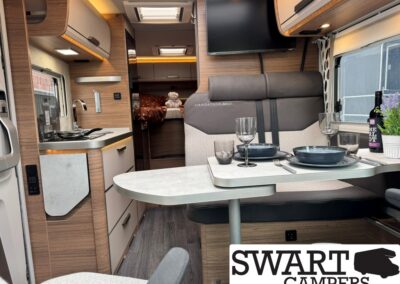 Interior of a modern camper van with a two-person dining nook, grey upholstered bench seating, and a small kitchen on the left; a bed is visible in the rear. A TV is mounted above the seating area and a window brings in light.