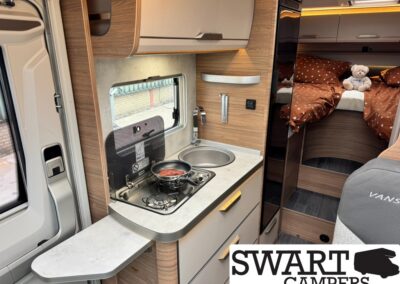 Campervan kitchenette with two-burner stove, pan, and sink, wood cabinets, and a bed with brown polka-dot bedding in the rear.