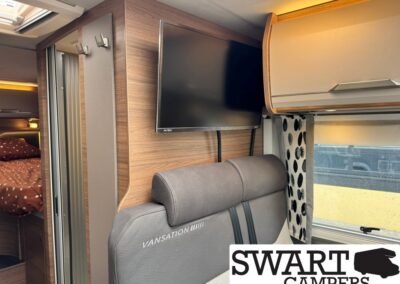 Interior of a Swart Campers van showing a wall-mounted flat-screen TV above a gray bench seat, wood paneling, a window with patterned curtains, and a bed with brown polka-dot bedding visible to the left.