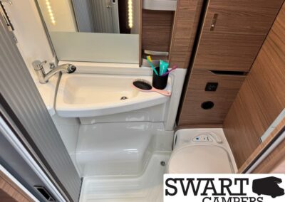 Compact caravan bathroom with white sink, chrome faucet, mirror, and toothbrush cup on a wood cabinet next to a small toilet.