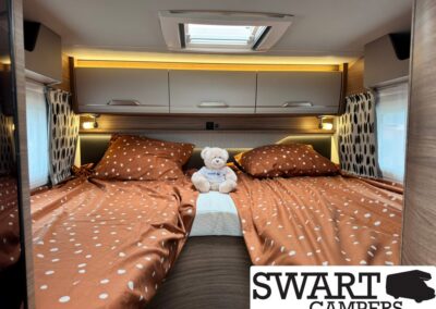 Rear bedroom of a campervan with orange polka-dot bedding, two pillows, a white teddy bear in the center, and overhead cabinets.