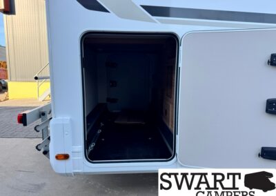 Open rear storage compartment of a white camper, showing a dark interior with a wooden cabinet on the right and a small board on the floor; logo 'SWART CAMPERS' partially visible at bottom right.