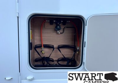 Open storage compartment of a camper with red propane hoses, a regulator, and black cargo straps.