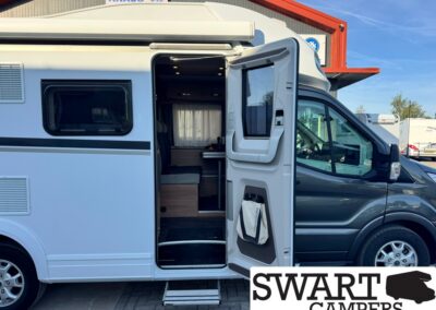White camper van with its side door open, exposing a small living area and window, parked at a dealership site with SWART CAMPERS/logo nearby.