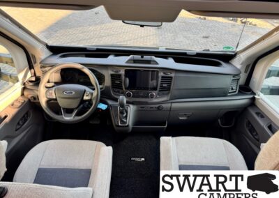 Interior view of a Ford camper van: steering wheel, center touchscreen, gear shifter, and grey front seats as seen from the back seat area.