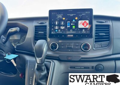 Car interior with a large center touchscreen showing apps and navigation, gear shifter in front, and keys in the ignition beside the steering wheel.