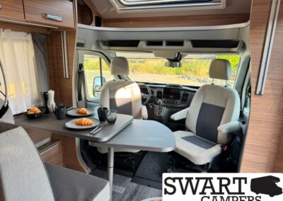 Interior of a campervan: a small dining table set for two with croissants, plates and mugs; front seats visible, SWART CAMPERS logo bottom-right.