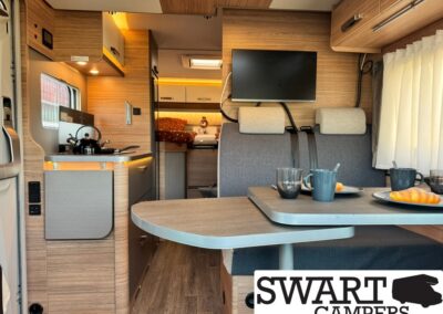Camper van interior with a small kitchen, table setup, and gray upholstered seating; TV on wall and a bed visible toward the back.
