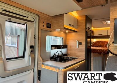 Camper van interior with a compact kitchen: two-burner stove, kettle, and wood-toned cabinets, plus a bedroom area in the back.