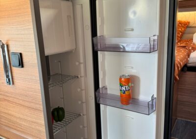 Open compact refrigerator in a camper van with a single Fanta can on the middle door shelf; pepper visible on bottom left shelf.