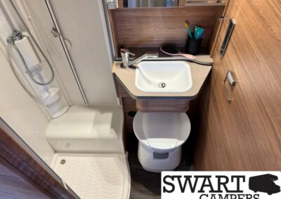 Compact camper bathroom featuring a small sink with faucet, toilet, and corner shower stall with wood-panel walls and storage shelf above.