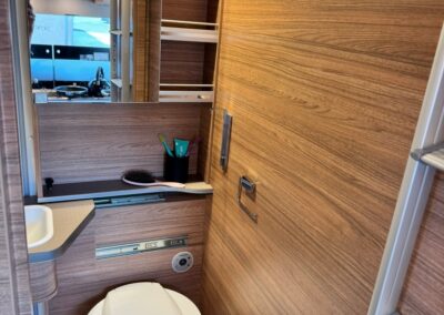 Small camper bathroom with wooden panel walls, a compact toilet, sink, and mirrored cabinet above.