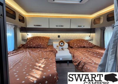 Camper bedroom with two single beds in brown polka-dot bedding, a small plush lion between them, overhead cabinets, and warm lighting under a skylight.
