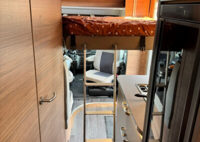 Interior of a camper van with a loft bed reached by a metal ladder, kitchenette on the right, wood cabinetry, and a front cab visible through the aisle, Swart Campers logo at bottom right.