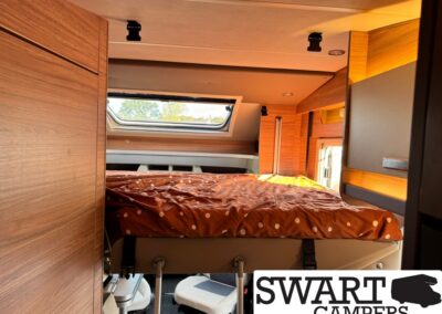 Interior of a camper van bunk bed area with wood paneling, brown polka-dot duvet, and built-in storage under a skylight window.