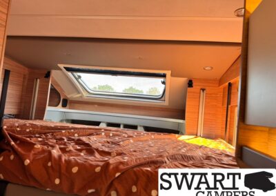 Cozy camper bedroom with a polka-dot brown duvet on a low bed, wood paneling, and a skylight letting in sunlight, SWART CAMPERS logo at corner.