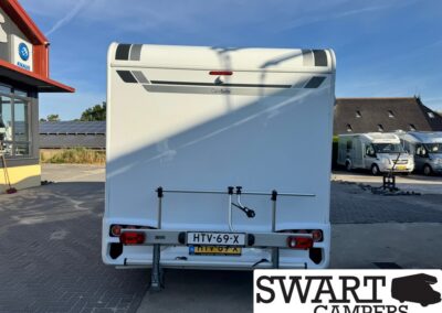 Rear view of a white caravan in a camper lot, with a bike rack and Dutch license plate HTV-69-X; SWART CAMPERS logo visible in corner.