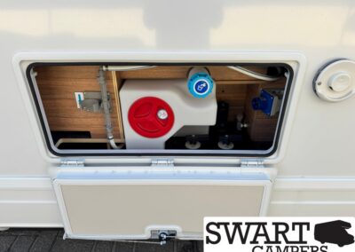Open camper service hatch showing water system components: white reservoir with red cap and blue fill cap, wood panel interior.