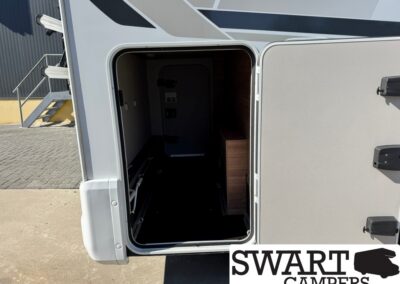 Open exterior storage compartment of a motorhome, revealing a dark interior and a wooden cabinet inside; SWART CAMPERS watermark visible.