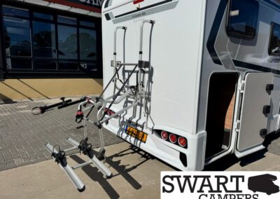 Rear view of a white camper with a mounted bike rack on the back, metal arms extending outward, and a side door open to the right; SWART CAMPERS logo is visible bottom right.
