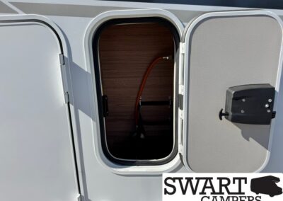 Open storage compartment on a camper trailer, showing a wooden interior and an orange flexible gas hose trailing inside.