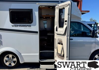 White camper van with the side door open and a lowered step, interior seating and kitchenette visible inside.