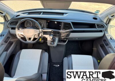 Interior of a modern van cabin: steering wheel, digital instrument cluster, center touchscreen, and gear shifter between two front seats; SWART CAMPERS watermark bottom right